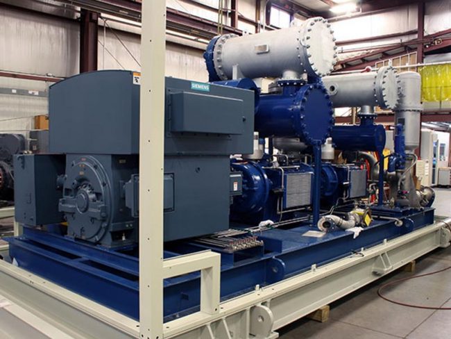 processing skid for oil and gas applications. A large industrial air compressor is positioned inside a warehouse. The machine features various pipes, valves, and electrical components. The setting includes metal beams and other equipment in the background, indicating an industrial environment.