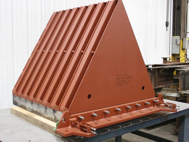 Image showing a large, industrial red metal fixture with a triangular shape, designed with multiple ridges. It is mounted on a flat metal base with bolts, placed on a workbench in a workshop. The background includes a white corrugated wall and other machinery elements.